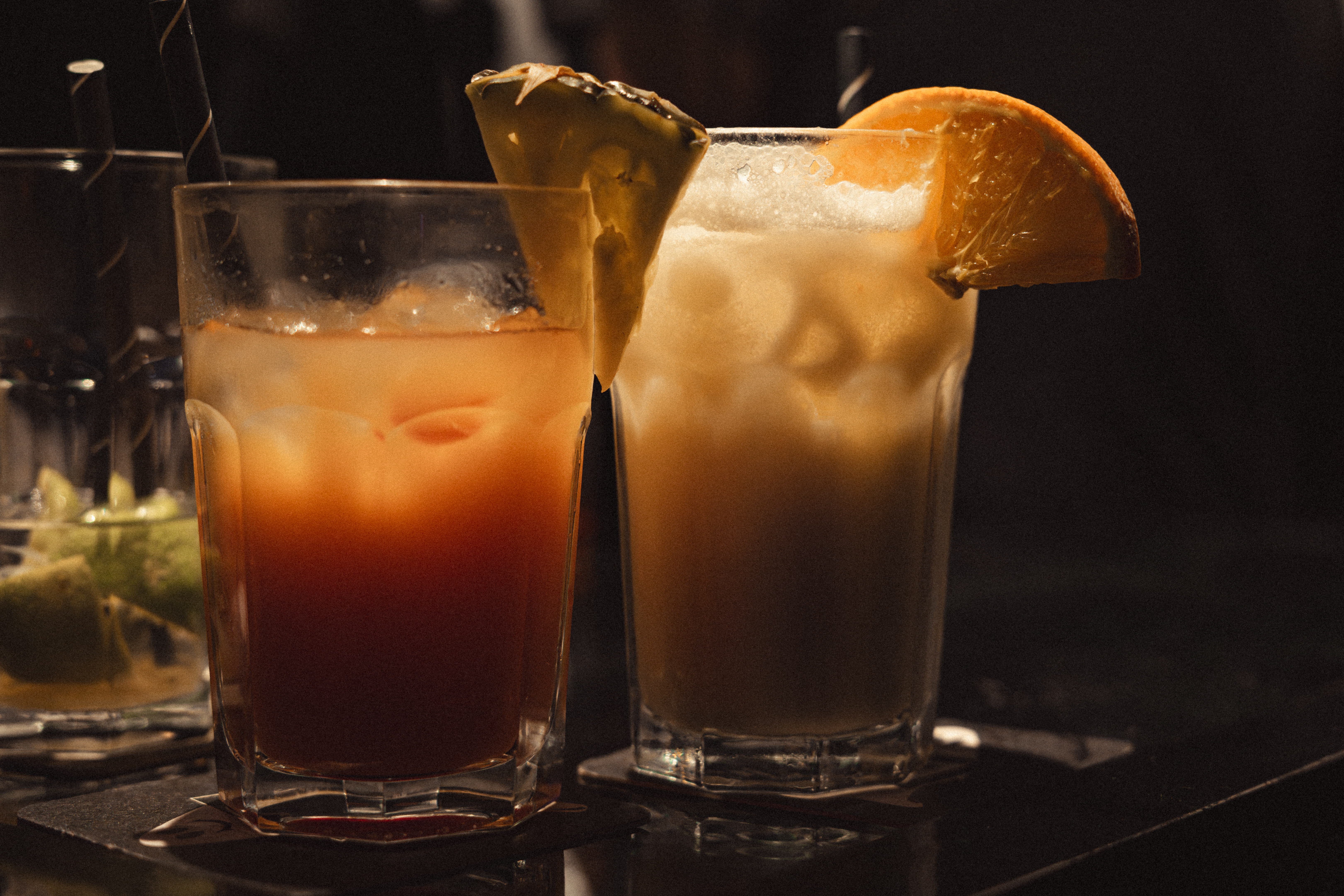 Drei Cocktails in Gläsern. Die zwei vorderen Cocktails sind noch voll und haben neben dem Strohhalm jeweils noch eine Ananas- und eine Orangenscheibe am Glas. Sie stehen auf einem schwarzen Glastisch.